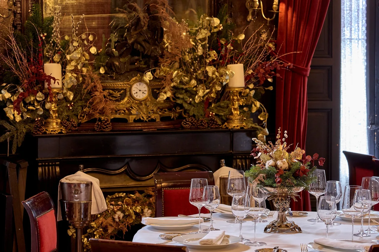An ornate dining room with gilded walls, red curtains and tables set with glasses and flowers.