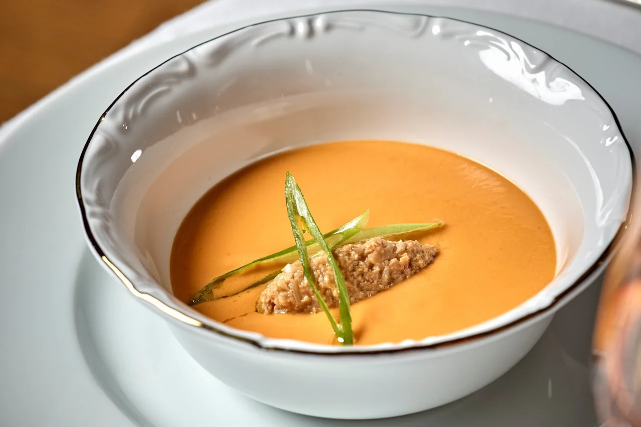 An orange cream soup with a small mound of shredded meat and green strips in a white bowl with a gold rim
