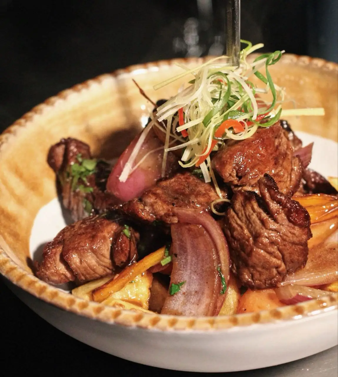 Lomo saltado of seared beef strips, red onion and peppers topped with shredded spring onion and chilli in a rustic bowl