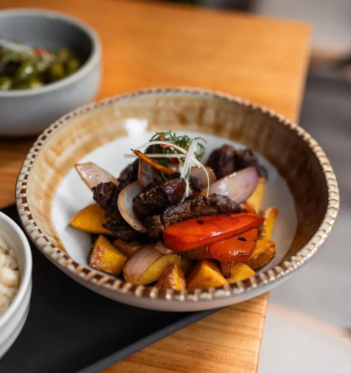 Bowl of lomo saltado with beef, onion, red pepper and fried potato wedges garnished with microgreens, beside a small rice bowl