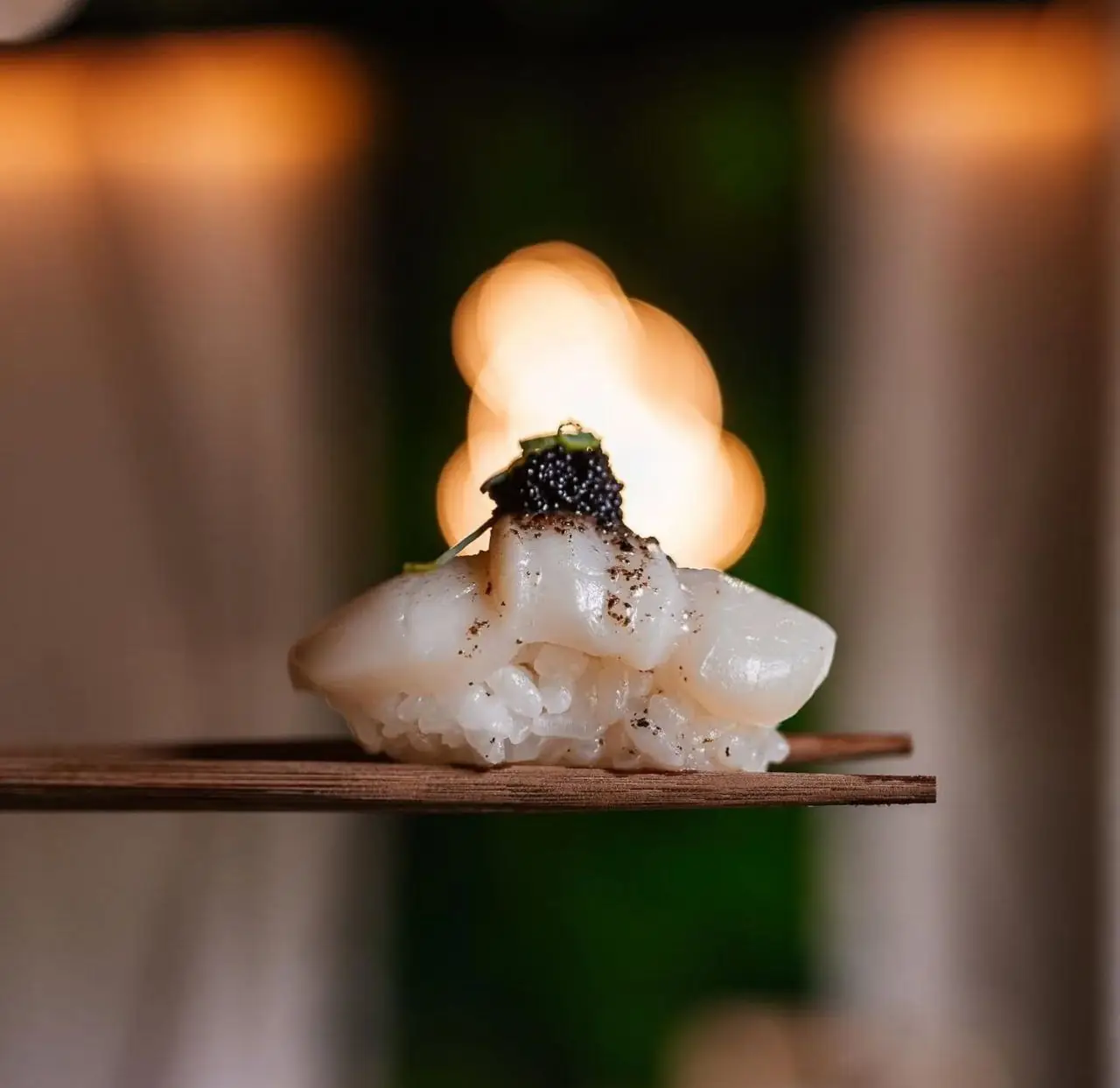 Single nigiri of pale seared fish topped with black caviar on a wooden board, warm bokeh lights behind