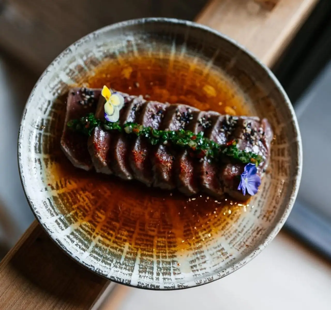 Overhead of thinly sliced seared beef tataki with sesame crust and a green herb line in amber ponzu, garnished with edible flowers in a speckled bowl