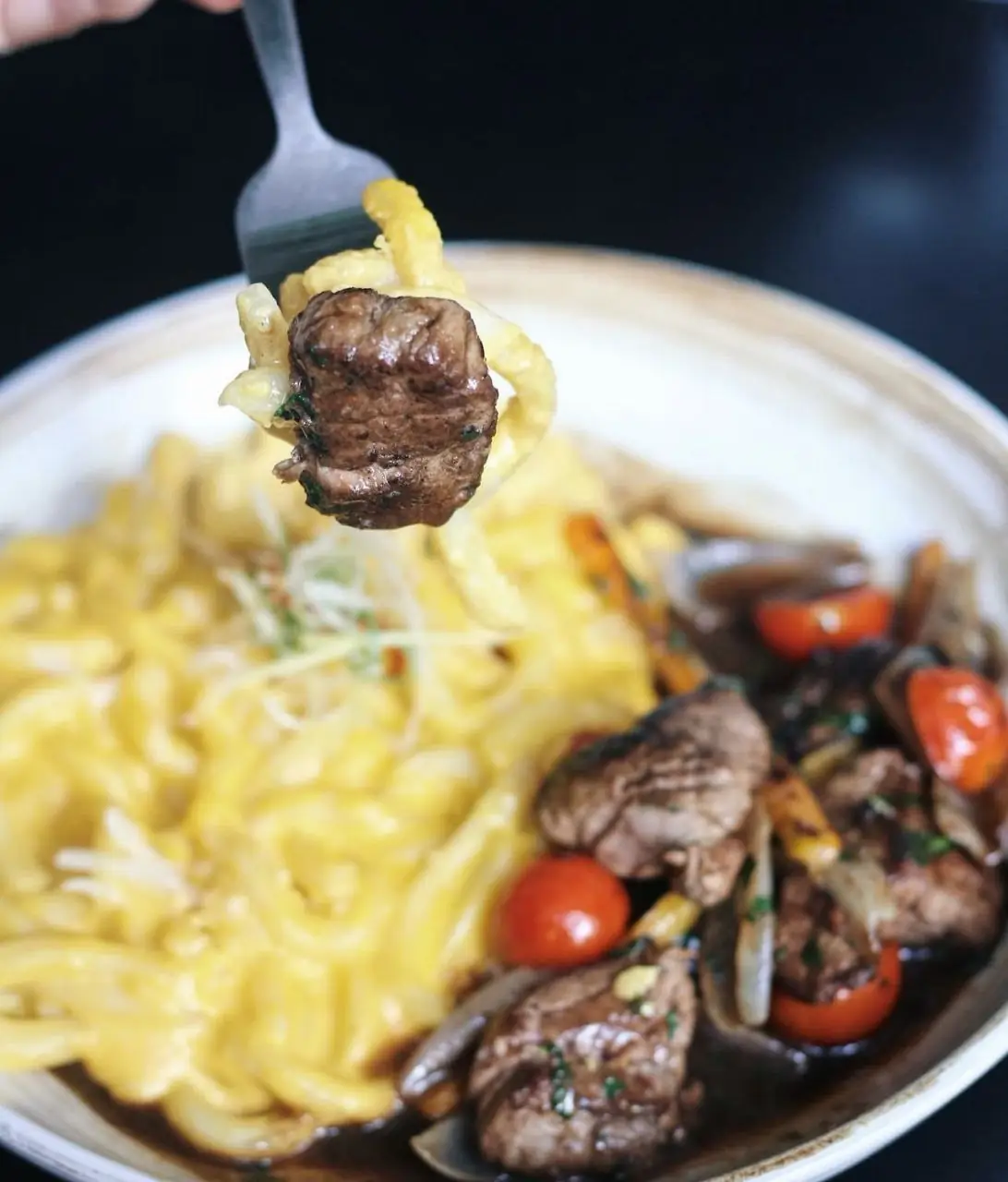 Plate of beef strips with cherry tomatoes and yellow noodles, a forkful of beef and noodles lifted above the dish
