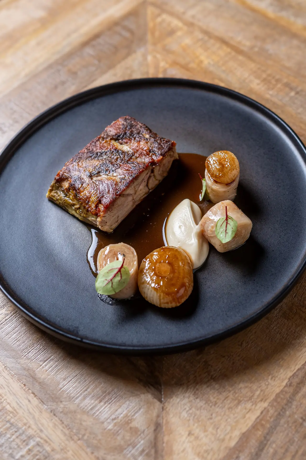 Crispy roasted pork belly with jus, glazed root vegetables and a purée on a dark plate