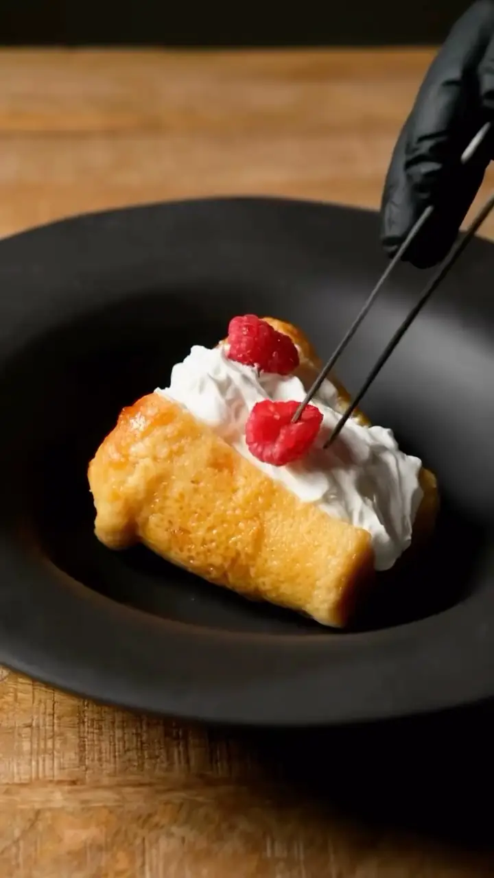 Hands plating a caramelized pastry with whipped cream and raspberries using tweezers