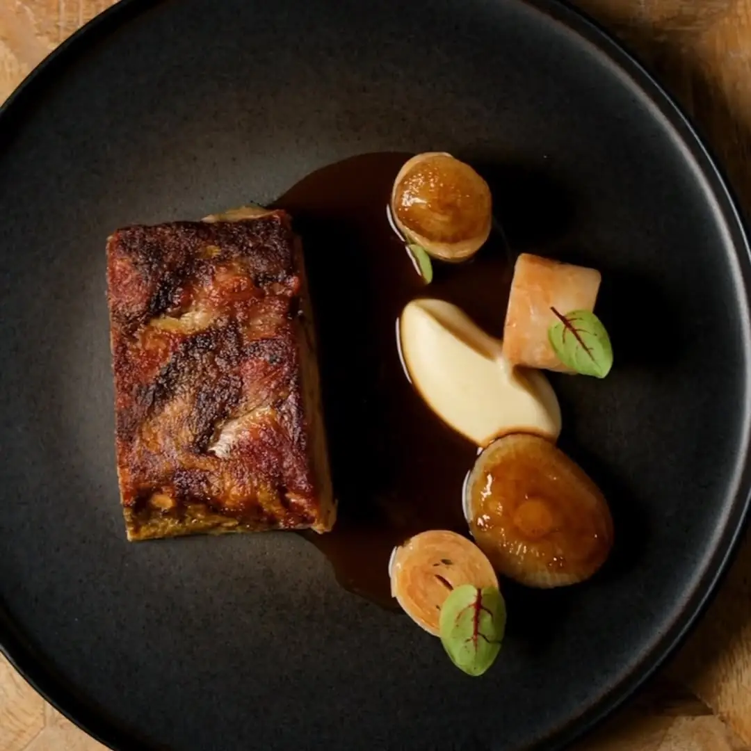 Seared pork belly with glazed shallots, apple and a light sauce on a dark plate
