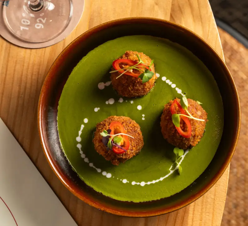 Three crispy breaded croquettes topped with sliced red chili and microgreens, plated over green sauce with dotted cream garnish, at Little Andaman