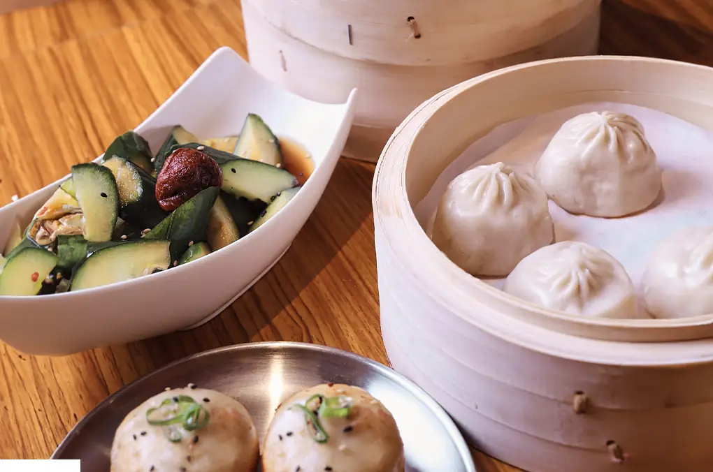 Bamboo steamer of soup dumplings beside a white bowl of smashed cucumber salad and a metal plate of pan-fried buns