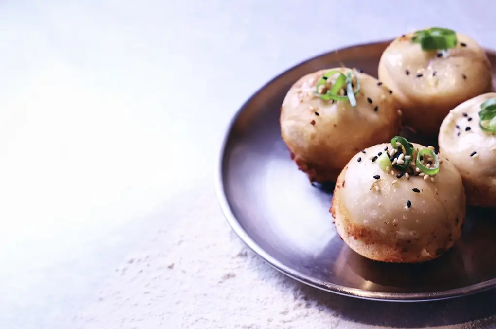 Four pan-fried buns topped with sesame seeds and sliced scallion on a metal plate over a floured surface