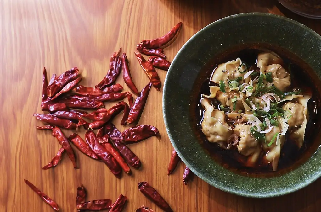 Green bowl of wontons in red chili oil with scallion and sesame, dried red chilies scattered on the wooden table beside it