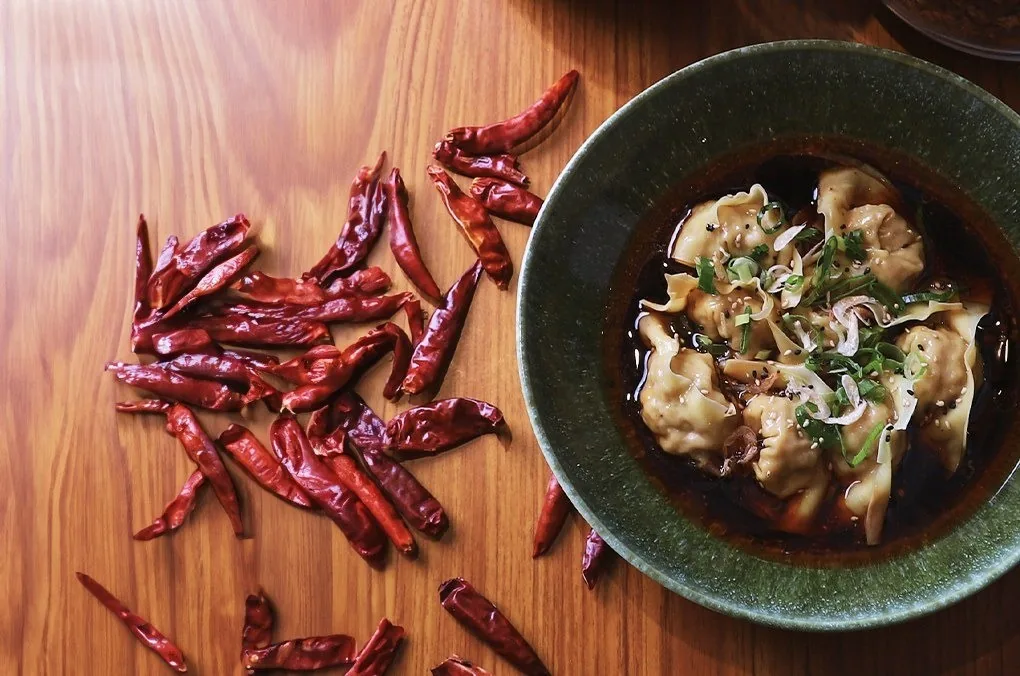 A green bowl of dumplings in dark chilli oil with dried red chillies scattered on a wooden table