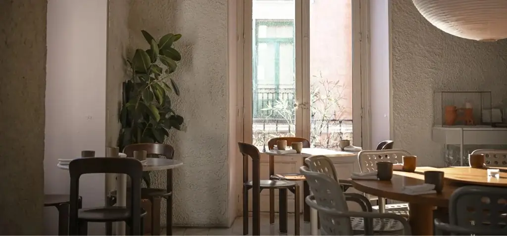 Warm-lit dining room with rough plaster walls, wooden tables and chairs, terracotta cups, a rubber plant and a tall balcony window