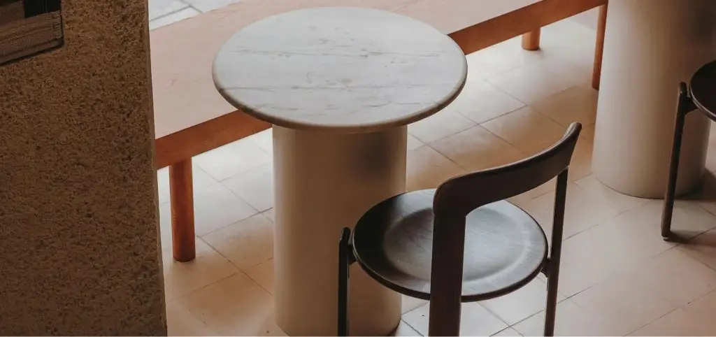 Detail of a round white marble pedestal table beside a dark wooden chair and bench on pale tiled floor