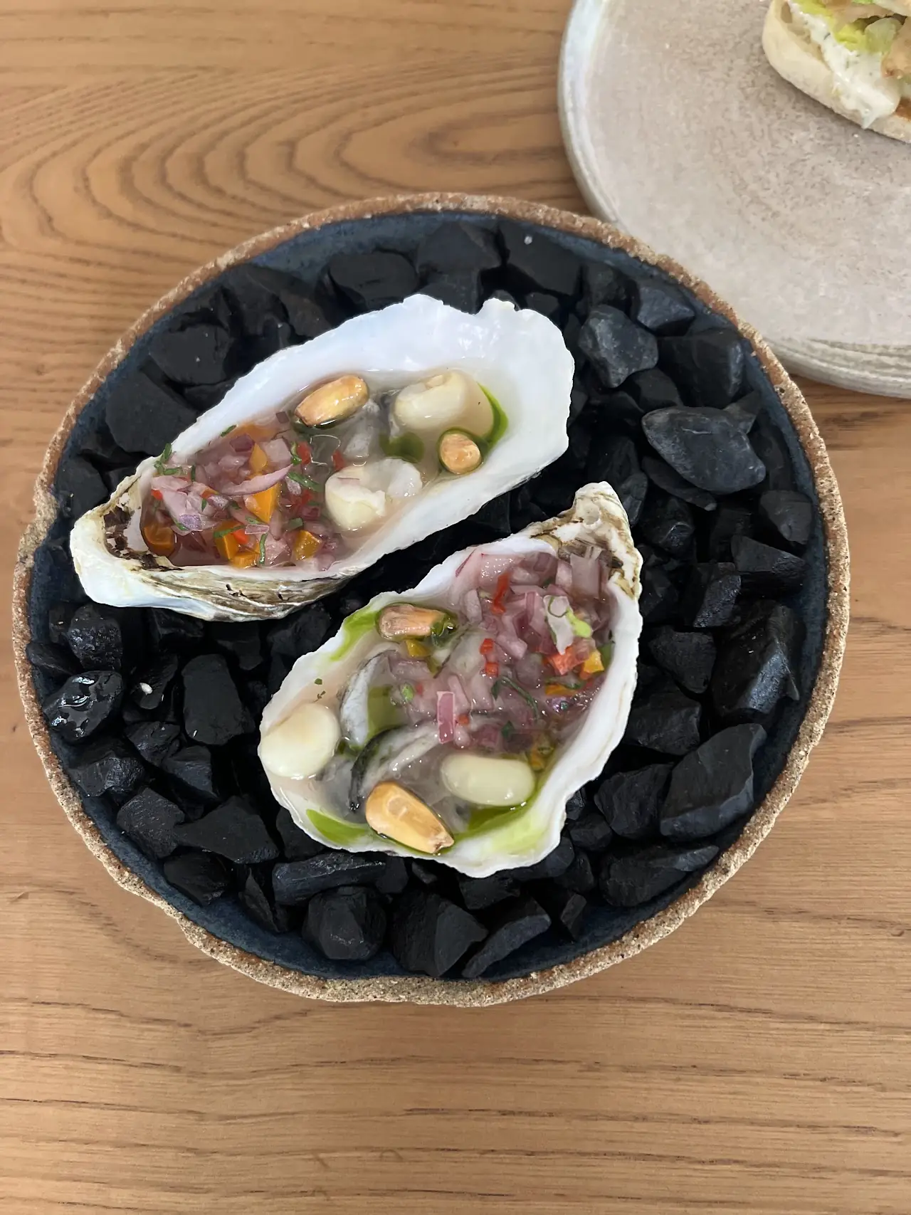 Two oysters topped with a ceviche-style garnish, served over black charcoal in a bowl.