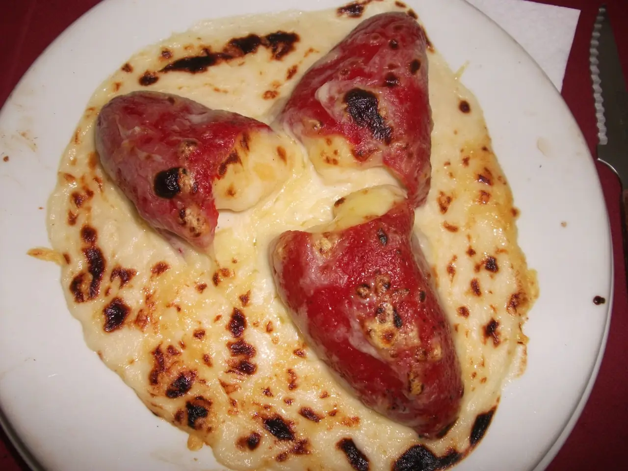 Three roasted red peppers stuffed and covered in melted, browned cheese sauce on a white plate at Restaurant La Llar de Foc