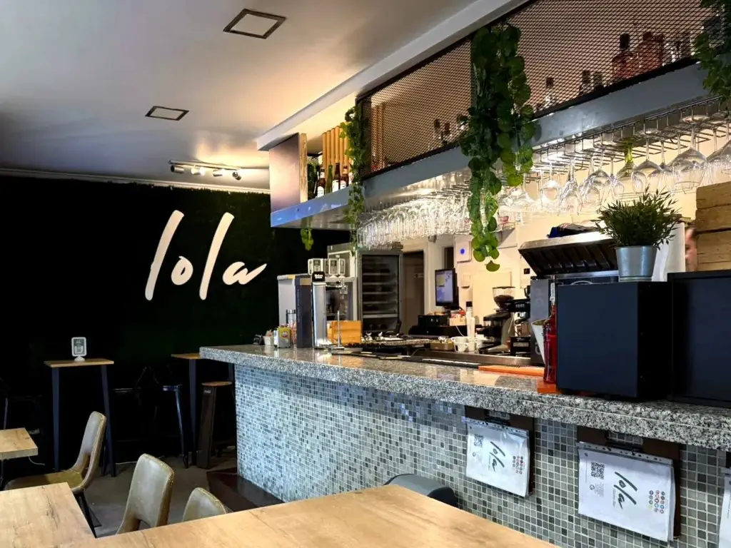 Interior bar with 'lola' painted on a dark green wall, mosaic-tiled counter, hanging wine glasses and trailing plants