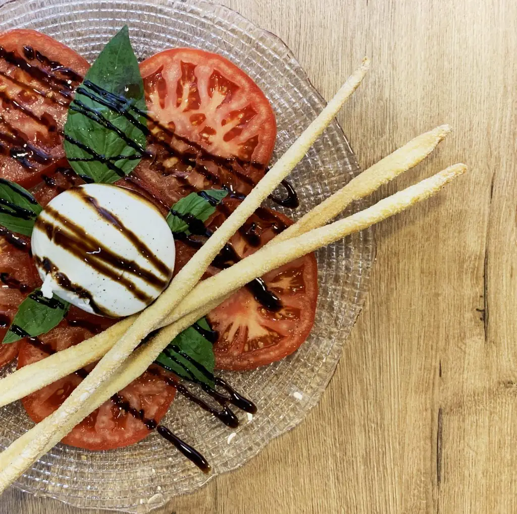 Caprese-style plate of sliced tomato, burrata, basil and balsamic drizzle with breadsticks on a glass plate