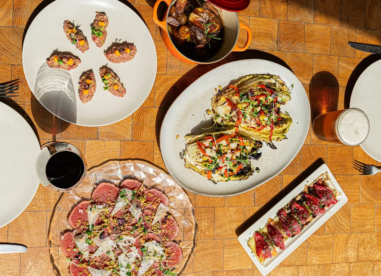 Overhead spread of beef tartare, sliced carpaccio with shaved cheese, grilled charred cabbage wedges with peppers, a skewer of seared meat bites, and a small pot of braised meat with potatoes on a wood table, at Lomo Alto