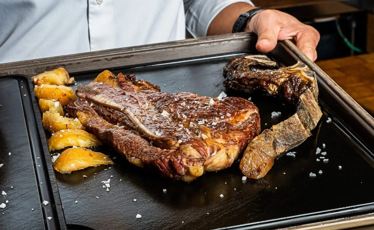 A cooked bone-in steak with charred crust and flaky salt, served alongside roasted potato wedges on a dark metal tray, at Lomo Alto