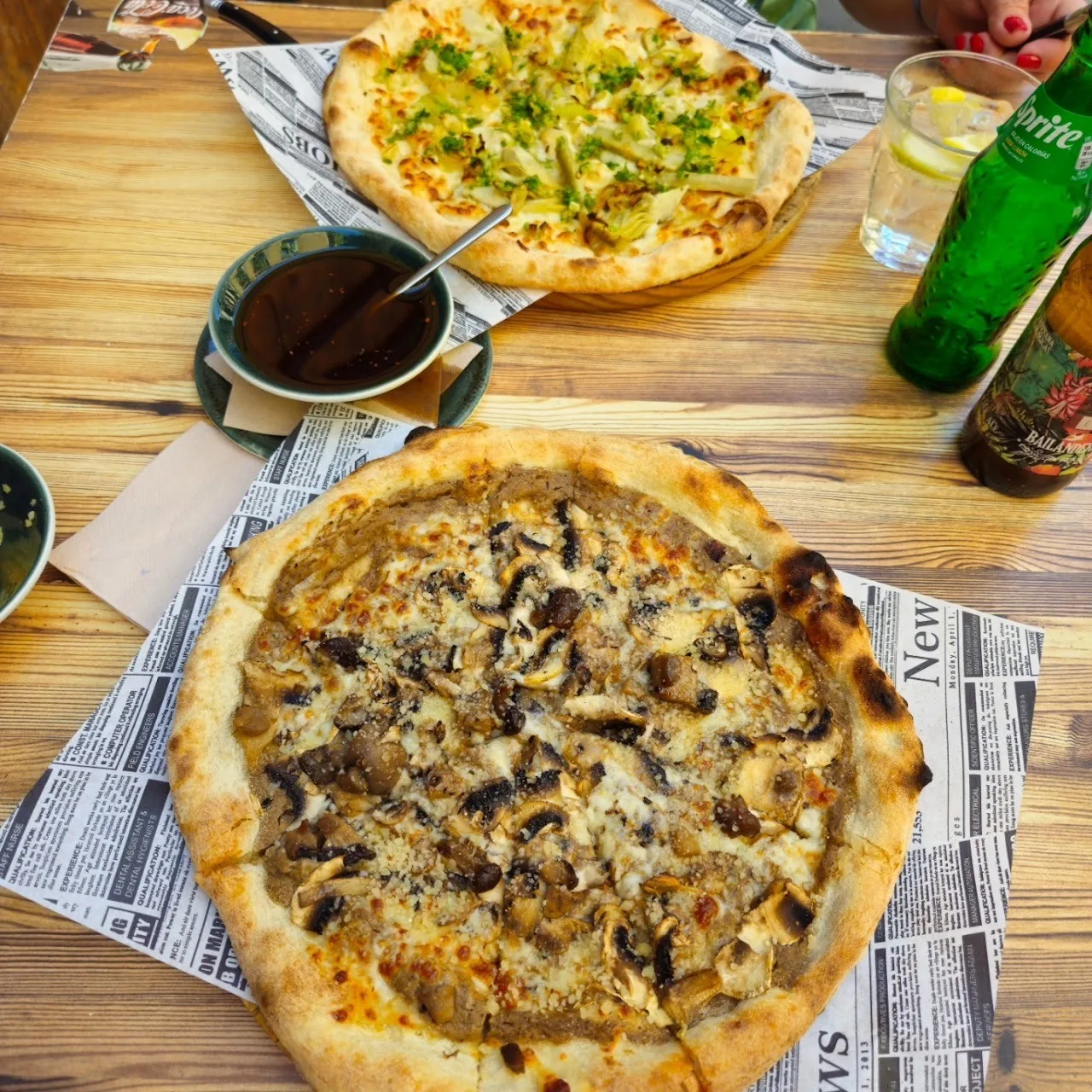 A pizza with dark mushroom toppings on a paper-lined table with a glass bottle beside it