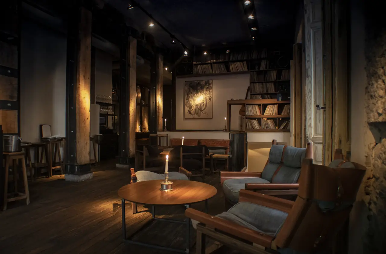 Candlelit lounge with leather and canvas armchairs around a round wooden table, shelves of vinyl records and a guitar along the back wall.