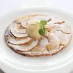 Thin apple tart dusted with icing sugar topped with a scoop of ice cream and mint on a white plate