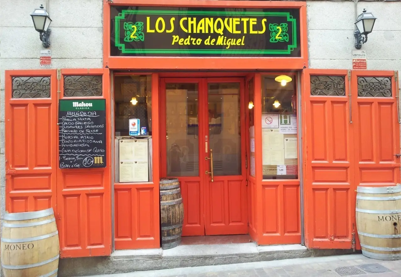 Storefront exterior of Los Chanquetes Pedro de Miguel with orange-red doors, a green-and-black sign, a chalkboard menu board, and two wine barrels flanking the entrance.