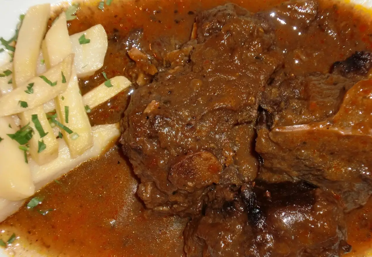 Close-up of a plate of braised meat in dark sauce next to sauteed potato strips topped with chopped parsley.