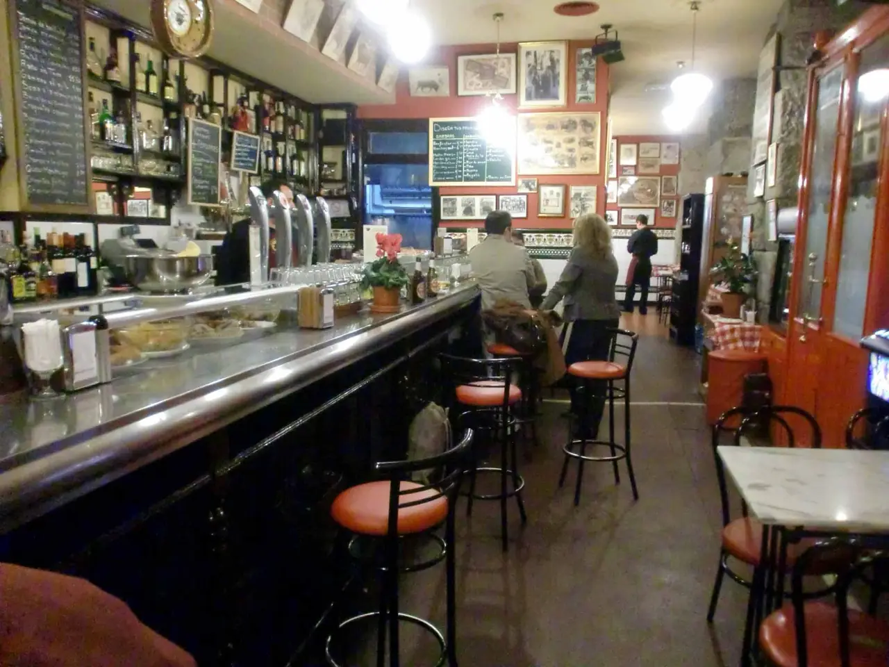 Narrow tiled bar interior with a long counter, beer taps, bottles on shelves, framed photos on the wall, red bar stools, and patrons standing near the back.