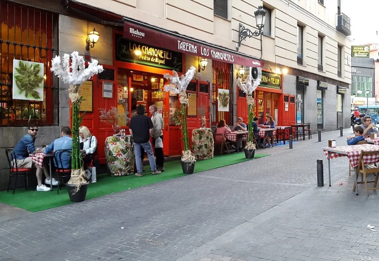 Street-level exterior of Taberna Los Chanquetes with red doors and an awning reading 'Taberna Los Chanquetes Pedro de Miguel,' diners seated at red-and-white checked outdoor tables on the sidewalk.