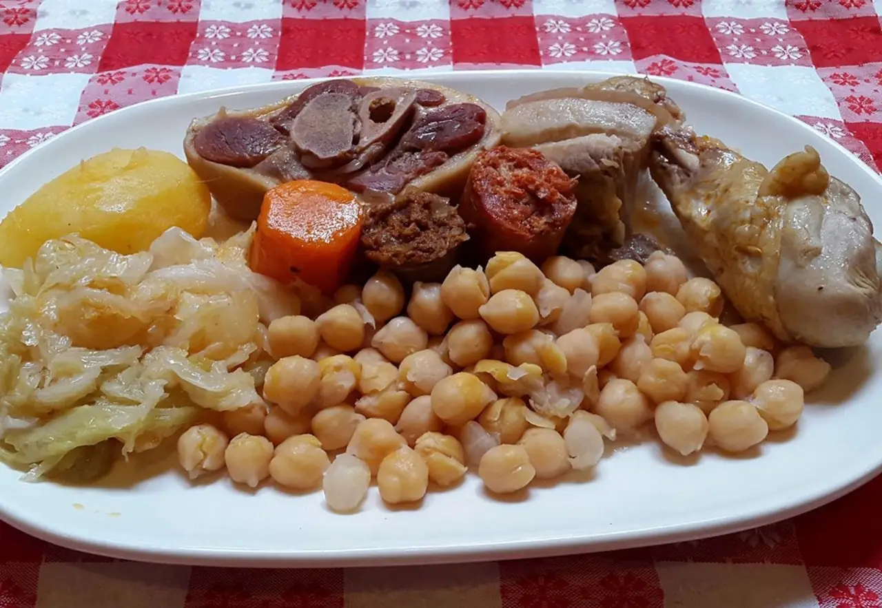 Oval plate of cocido madrileno with chickpeas, boiled cabbage, carrot, chorizo, morcilla, chicken and marrow bone, on a red-and-white checked tablecloth.