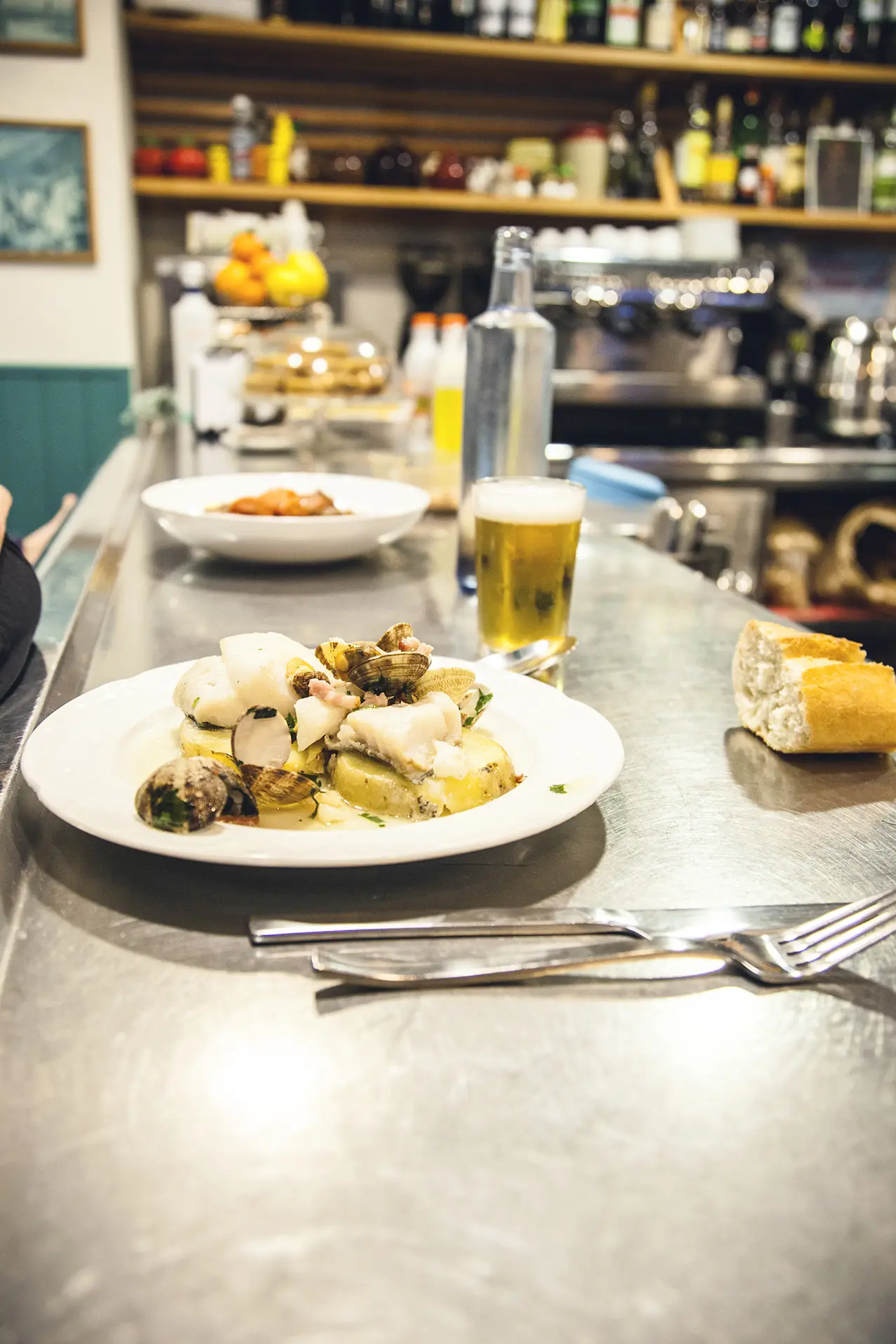 Plate of clams and white fish over sliced potatoes at the bar, with a glass of beer and bread