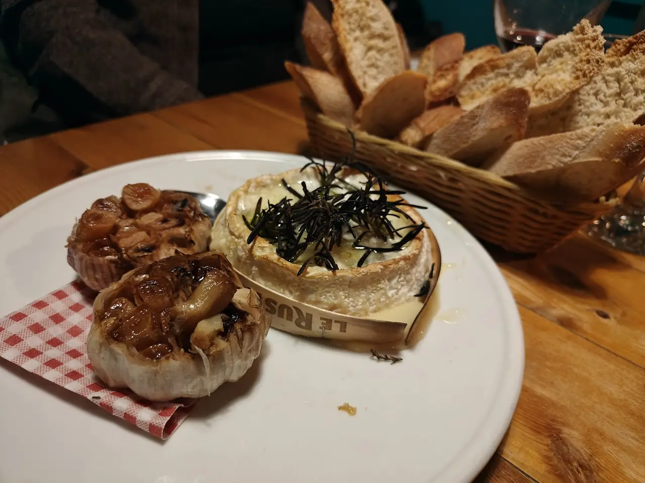 Baked whole cheese topped with rosemary next to roasted garlic bulbs and a bread basket