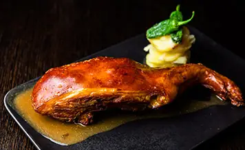 Quarter of crisp-skinned roast suckling pig in its own jus with mashed potato and a fried green pepper on a slate plate