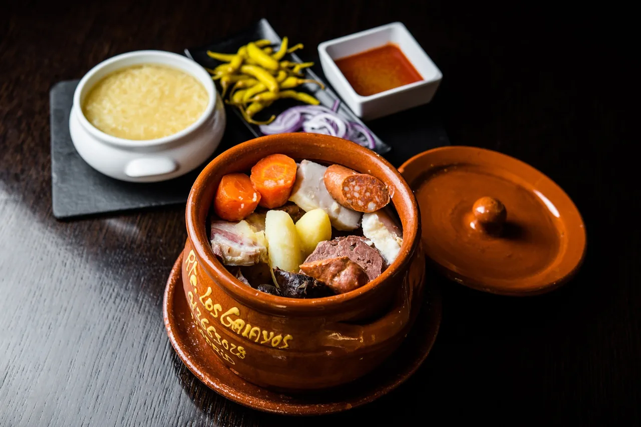 A clay pot of cocido with carrots, potatoes, chorizo and boiled meats beside a bowl of soup, green peppers and a small dish of sauce on a dark table