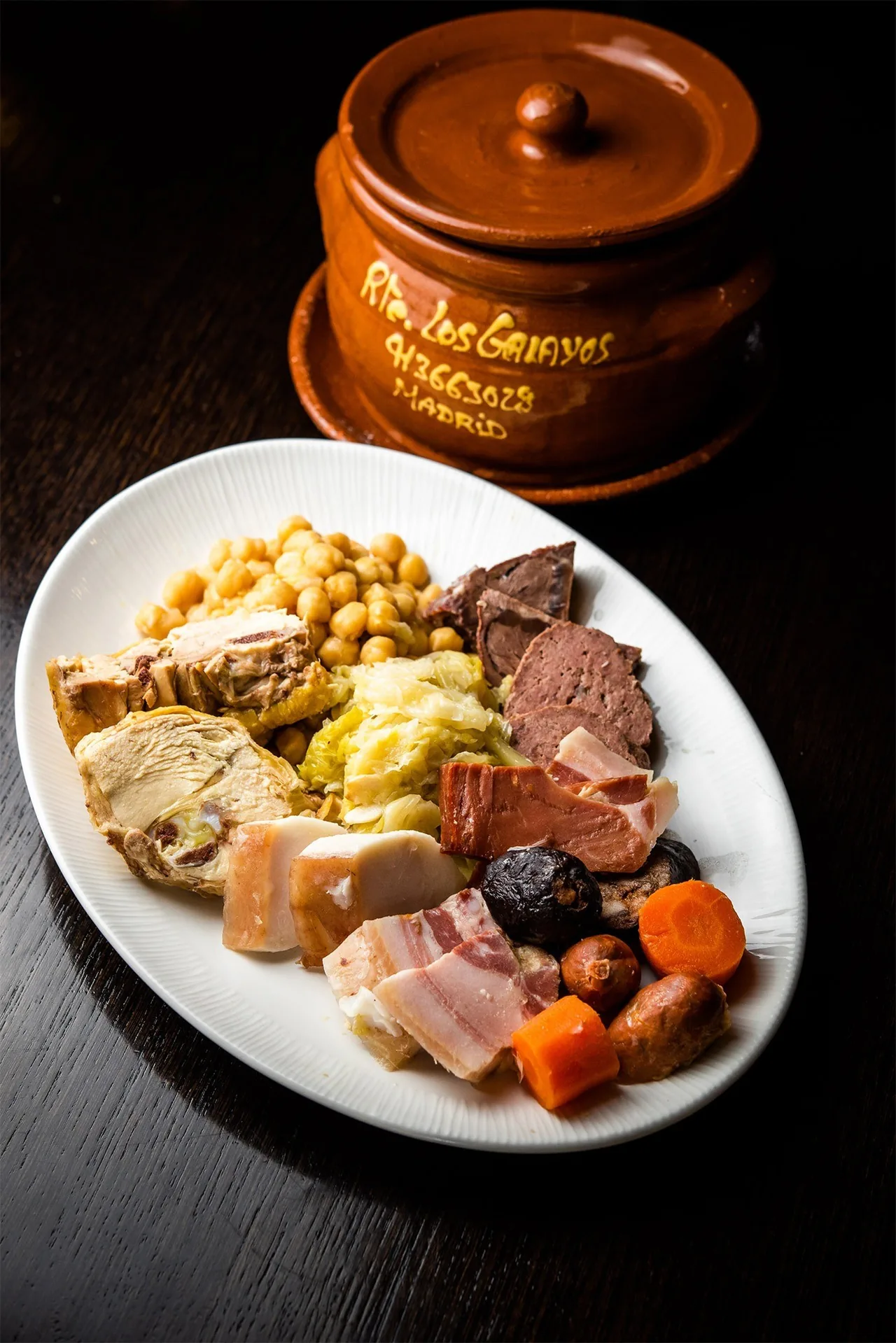 A white oval plate of chickpeas, cabbage, boiled meats, bacon and carrots beside a clay pot with its lid