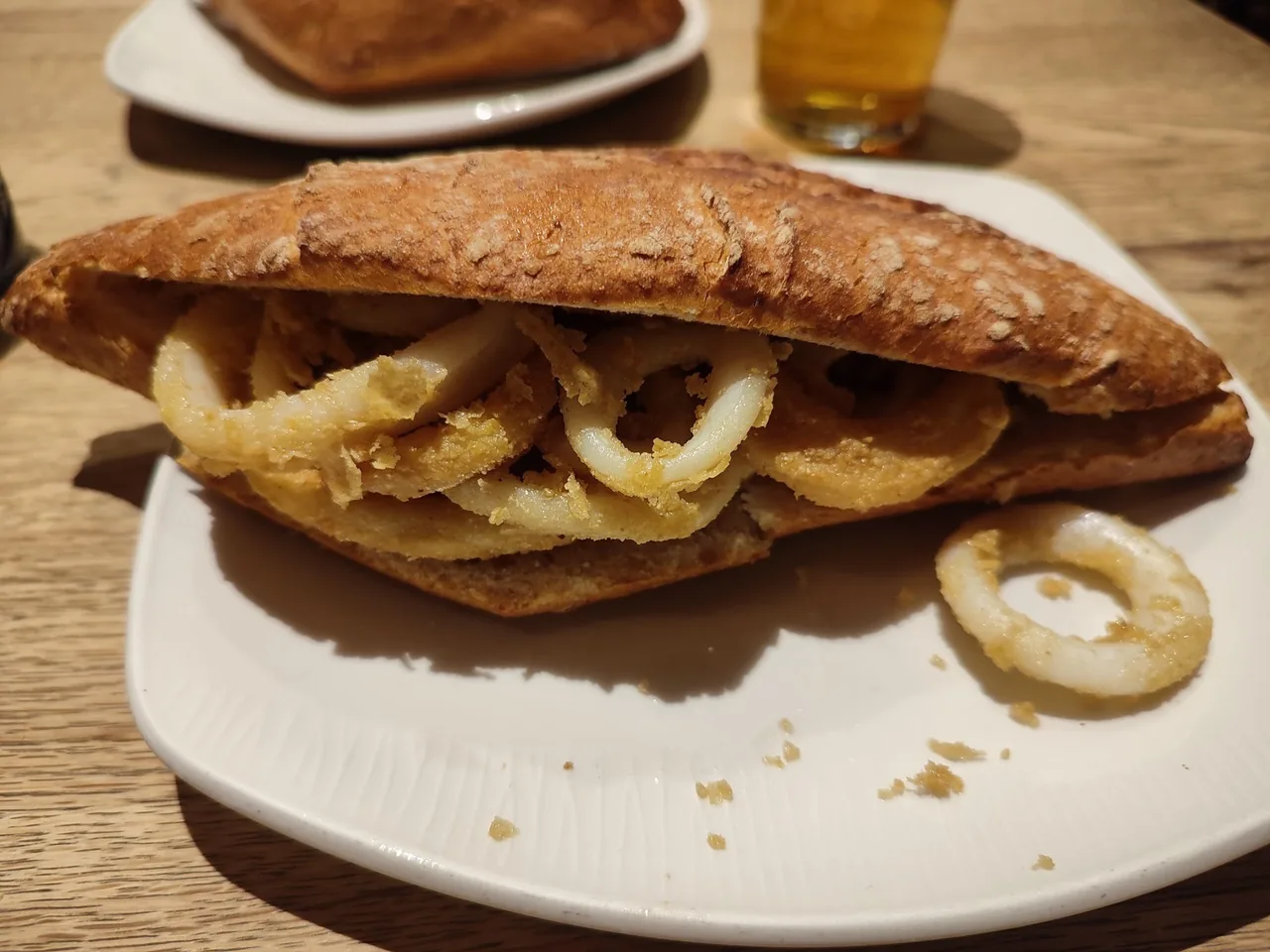 A crusty roll filled with fried squid rings on a white plate with a beer behind it