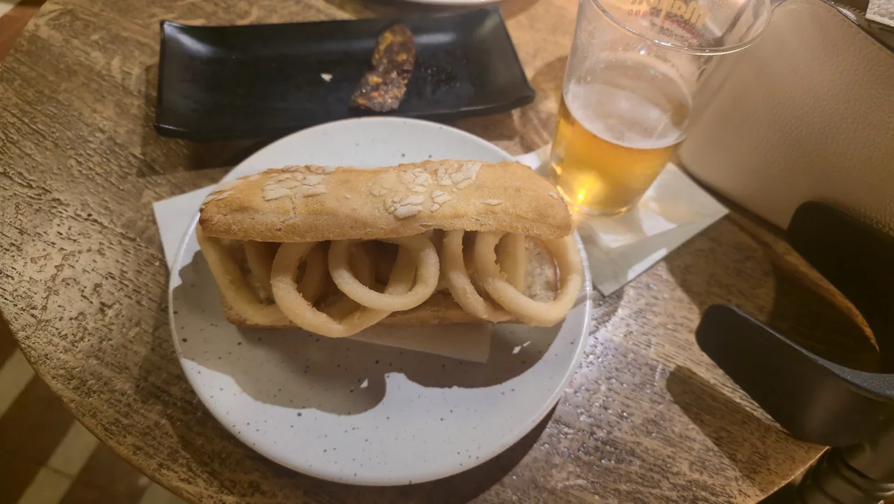 A roll filled with fried squid rings on a plate beside a glass of beer