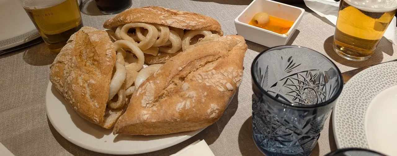 A crusty roll filled with fried squid rings on a white plate, with glasses of beer behind