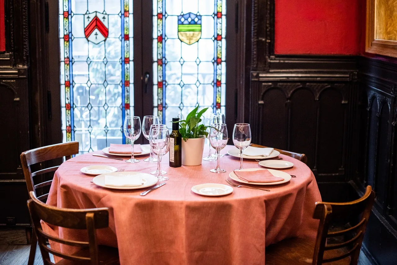 A round table set with a pink cloth beneath stained glass windows and dark wood panelling.