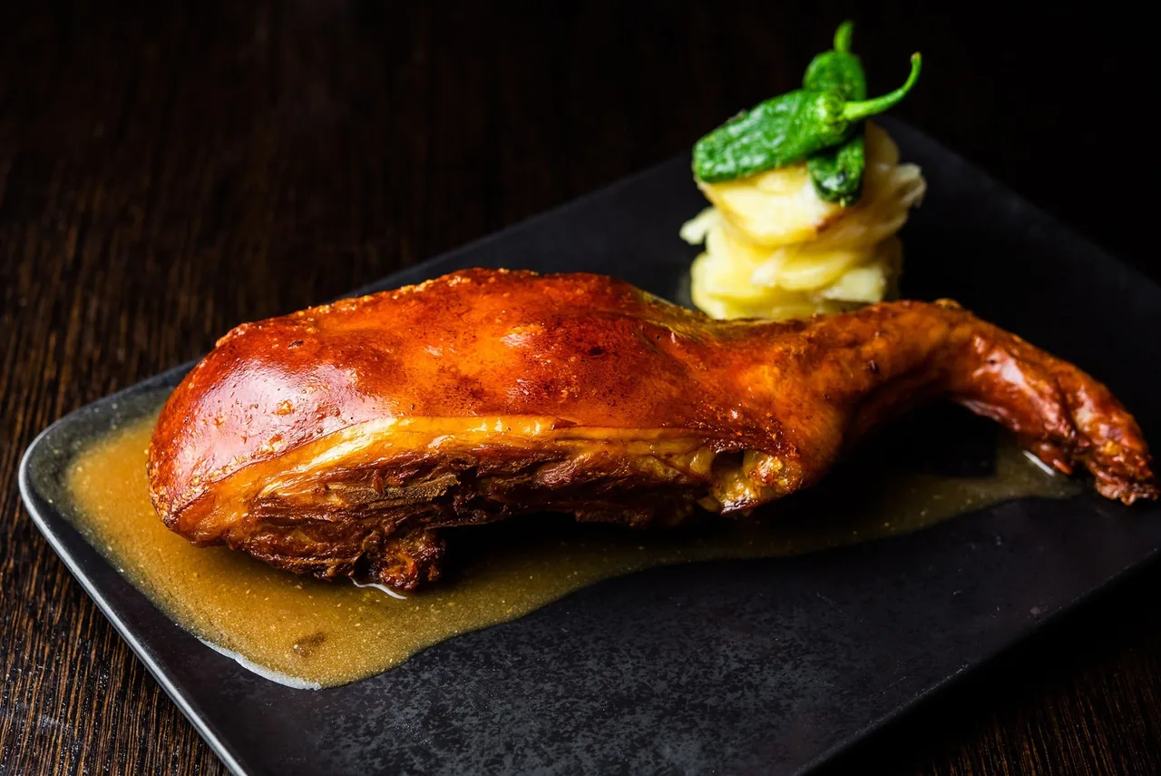 A roast poultry leg on a black plate with a garnish of potato and a green pepper