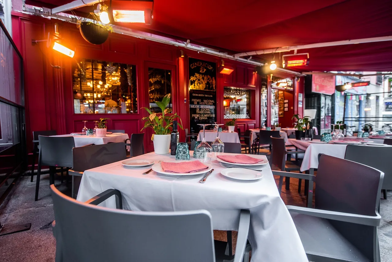 Tables laid with white cloths and pink napkins on a covered outdoor terrace beside a red facade