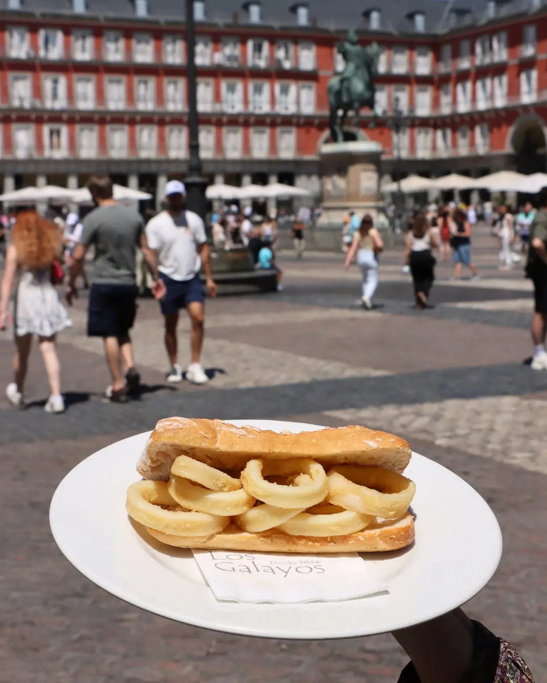 A baguette sandwich filled with fried calamari rings held up on a plate in a plaza