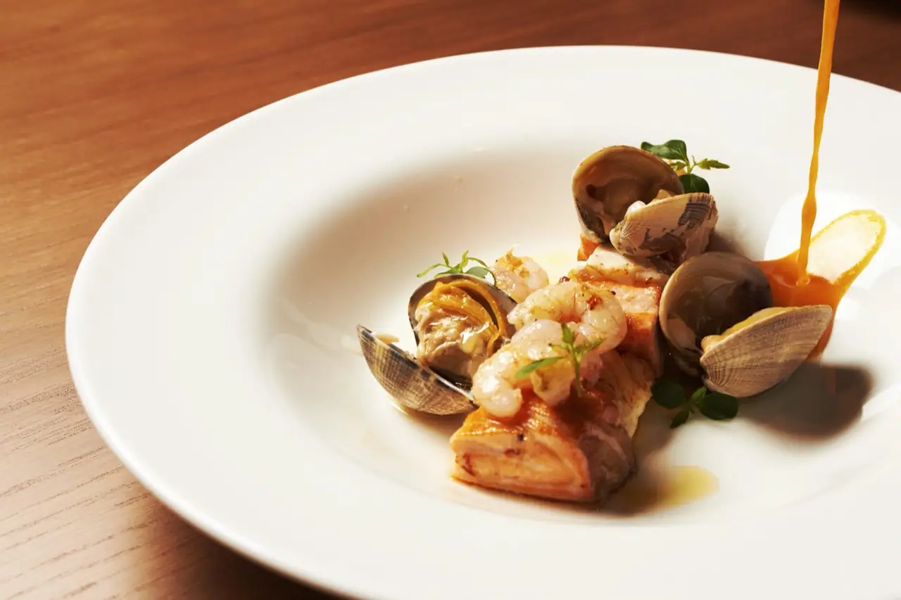 Seared fish fillet with clams and prawns on a white bowl as an orange sauce is poured over it