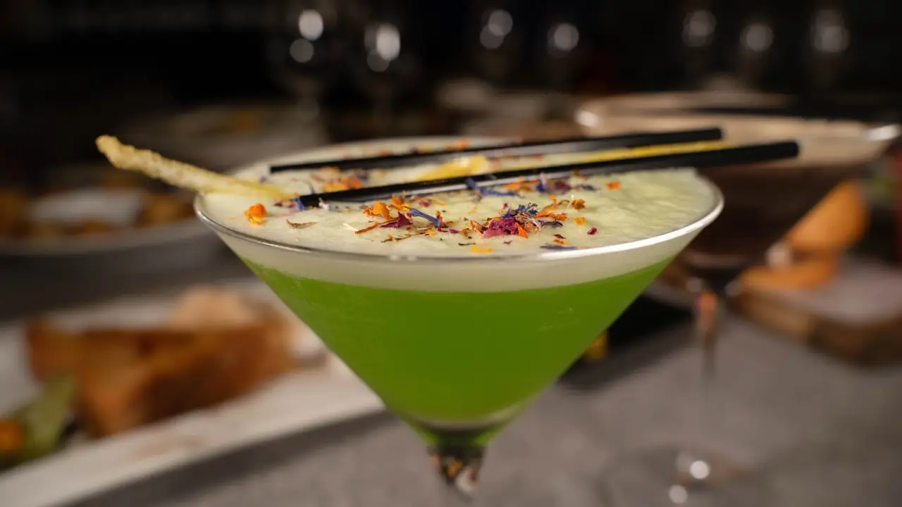 Bright green foamed cocktail in a martini glass topped with dried flower petals and a crisp garnish, black straws across the rim