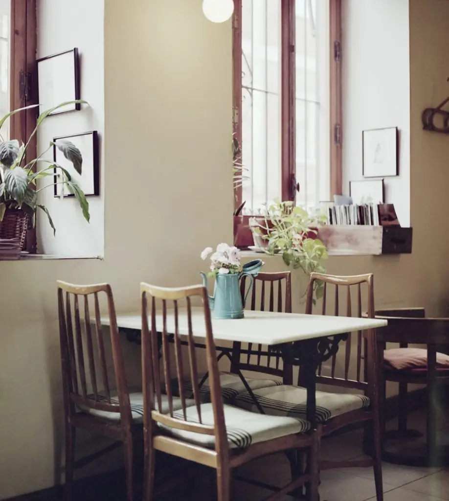 Cozy interior corner with vintage wooden spindle-back chairs around a marble-top iron table, a turquoise jug of flowers, framed botanical prints and potted plants by tall windows