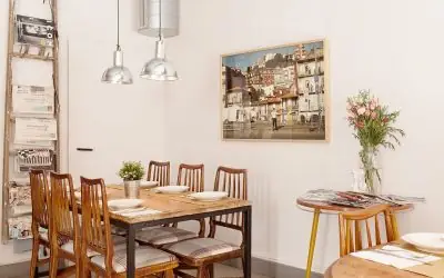Interior dining room with a long wooden table and spindle-back chairs, a framed cityscape painting, a magazine rack and a vase of pink lilies