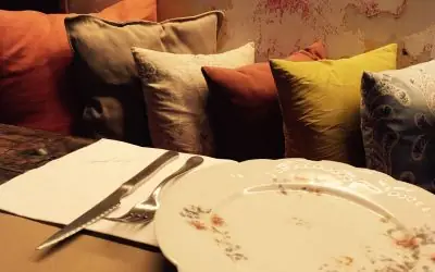 Interior detail of a set table with a floral-rimmed plate and cutlery beside a banquette piled with mismatched cushions