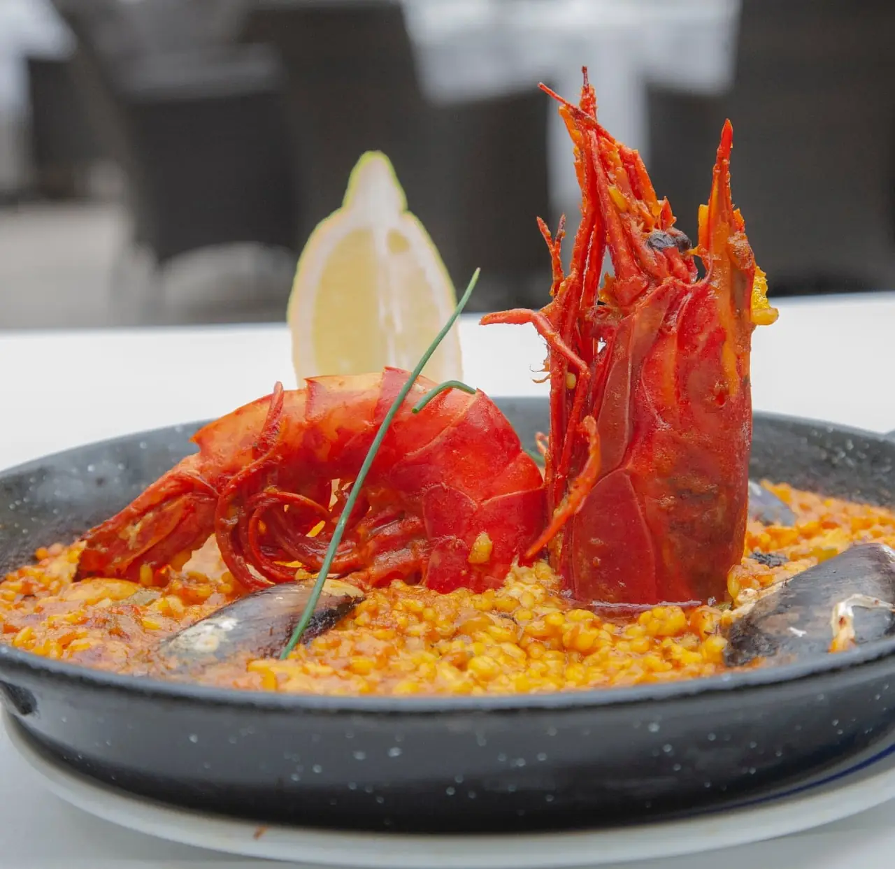 Paella pan topped with a whole grilled langoustine and mussels, garnished with a lemon wedge and chive
