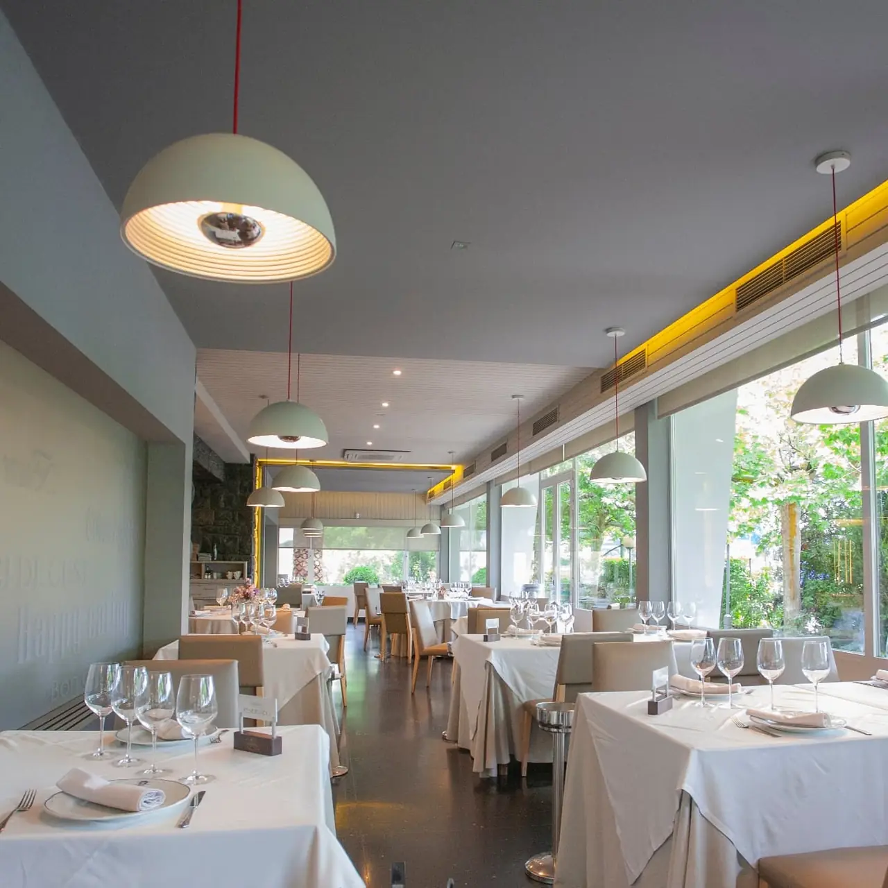 Bright dining room with white-clothed tables, wine glasses, and pendant lamps beside floor-to-ceiling windows overlooking greenery
